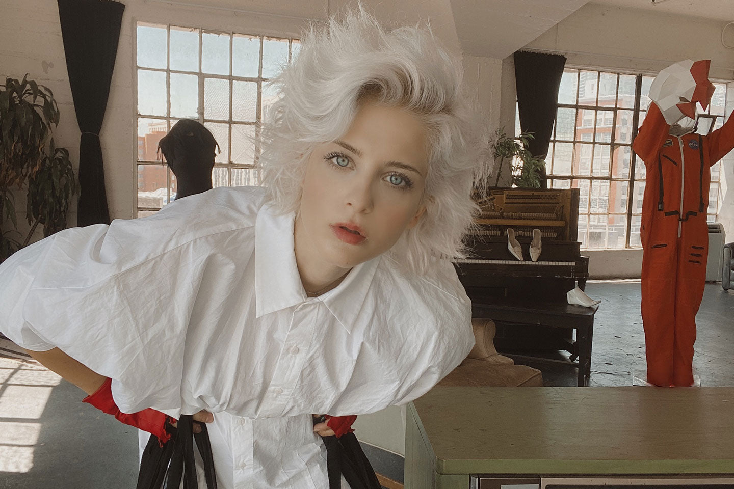 Photo of Buel with white hair and a white shirt leaning over and staring at the camera. there are large windows and a city skyline in the background.