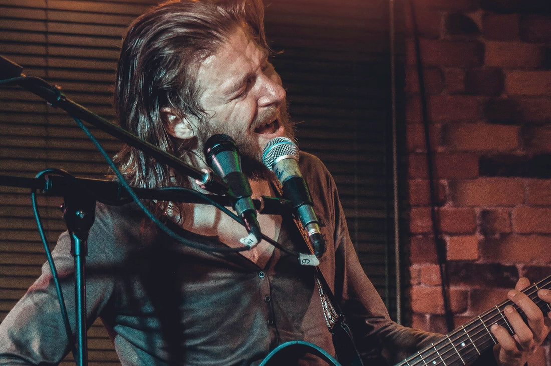 Photo of a bearded man with long hair playing guitar and singing into a microphone.