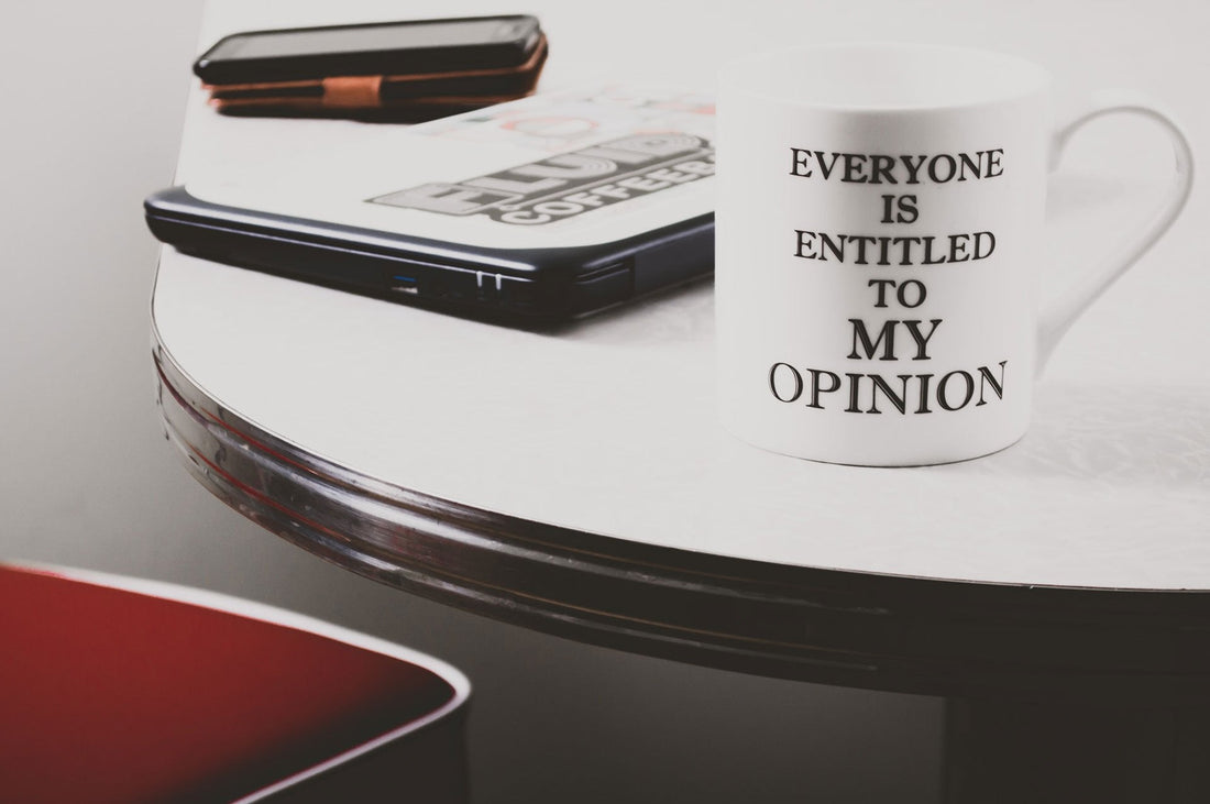 A photo of a white coffee cup on a white table. The Coffee cup says 'Everyone is Entitled to MY OPINION" on it in black letters