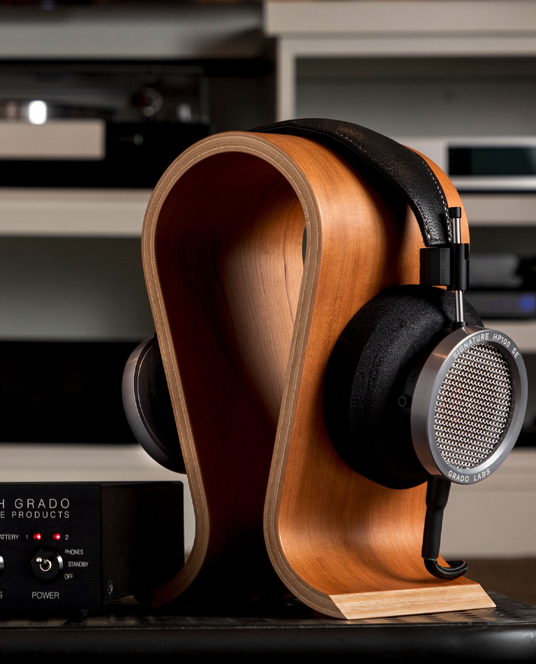 Photo of the HP100se headphones on a wooden stand infront of a white bookcase. The headphones are black with silver ear cups.