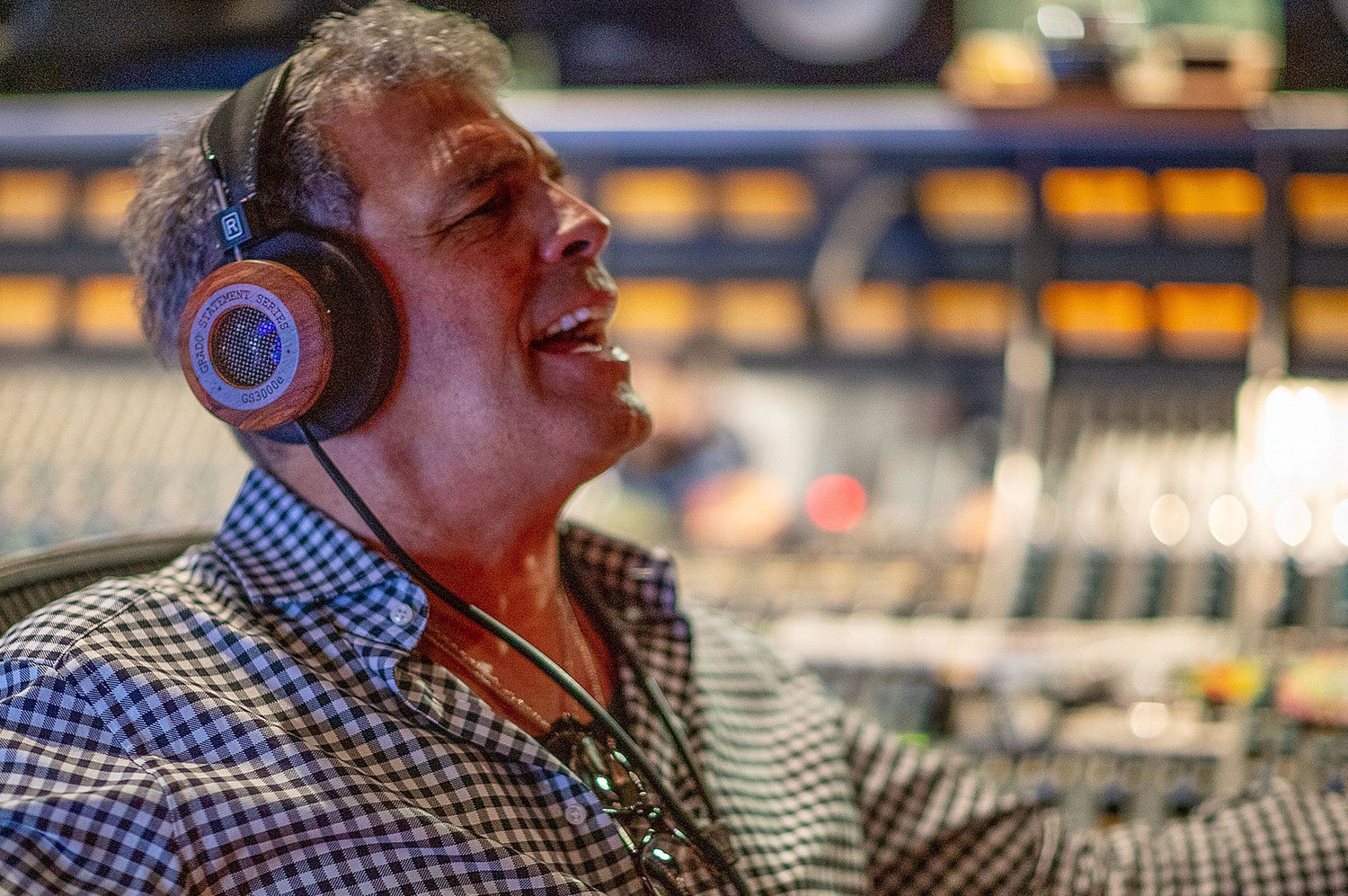 A photo of a man wearing a plaid shirt and wearing grado headphones while mixing music.