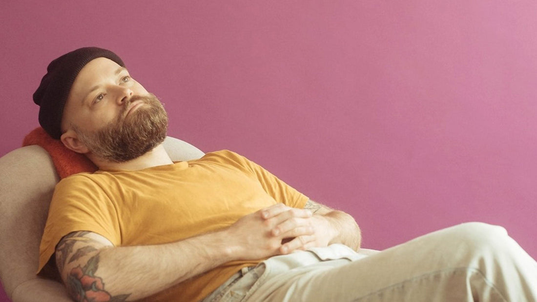 Photo of a man laying on a white chair in front of a pink wall. He has a red beard and is wearing a black beanie, a yellow t-shirt and white jeans.