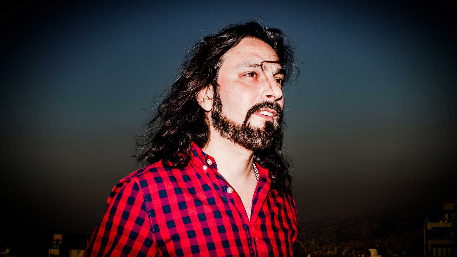 Photo of a man with a black beard and long hair. He is wearing a black and red checkered shirt and standing in front of a twighlight sunset.