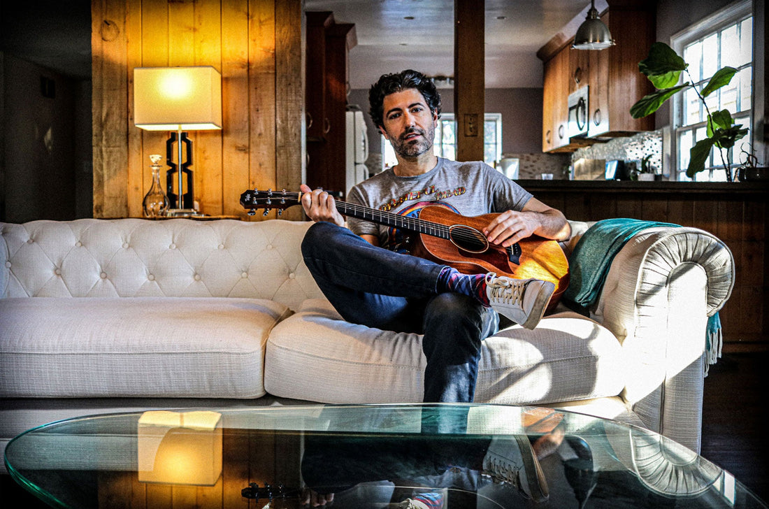 Photo of a man in blue jeans and a grey t-shirt playing an acoustic guitar while sitting on a white couch.