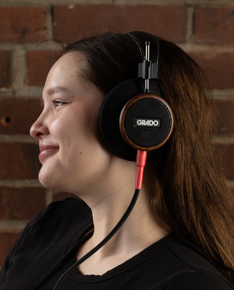  Photo taken from the side of a young black haired woman wearing grado s550 headphones in front of a brick wall.