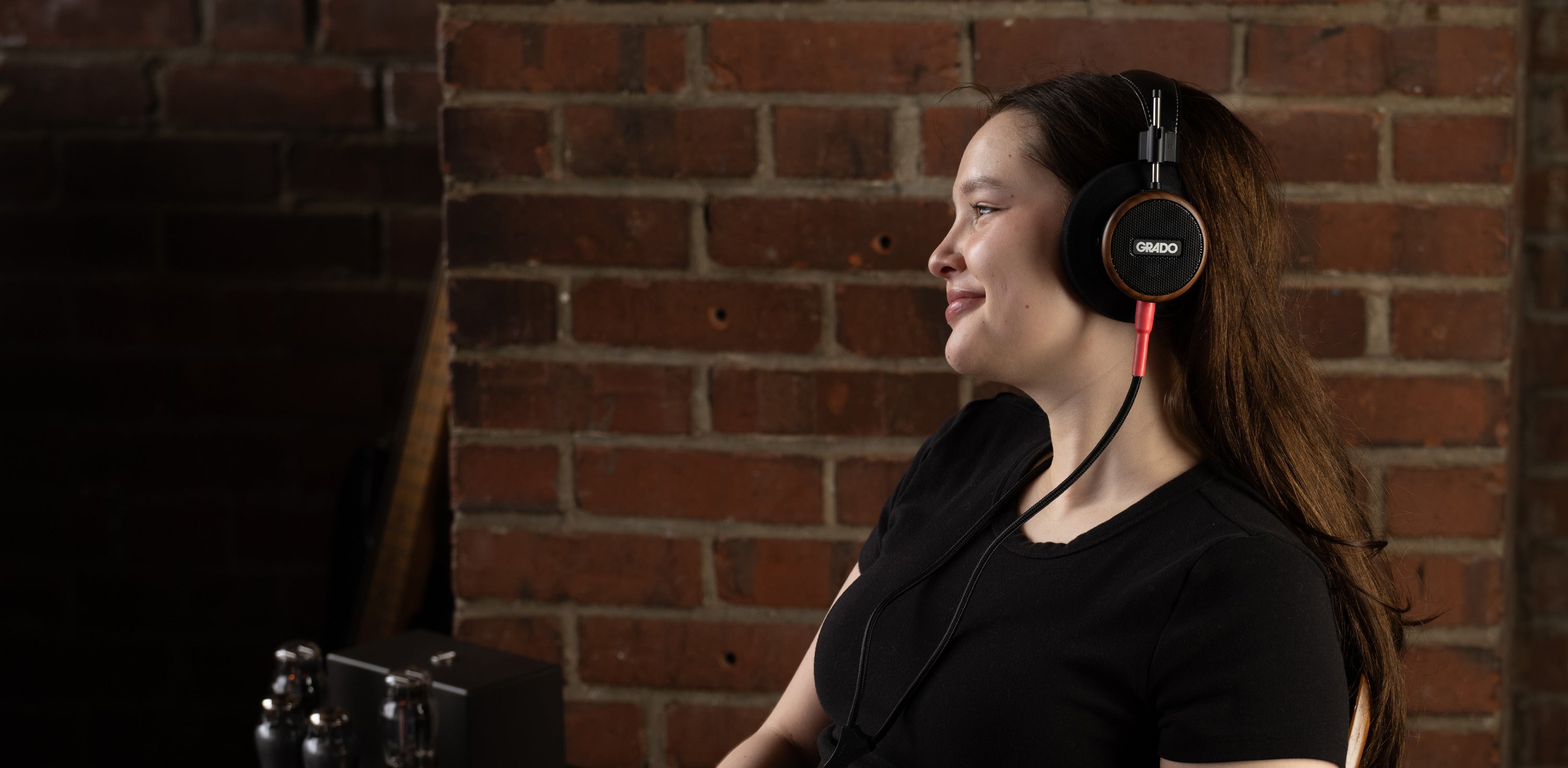  Photo taken from the side of a young black haired woman wearing grado s550 headphones in front of a brick wall.